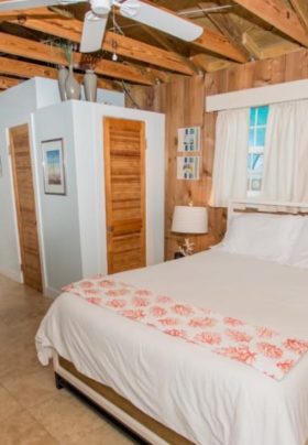 Rustic interior cottage view of bed underneath windows and open beam ceiling with kitchen visible in background